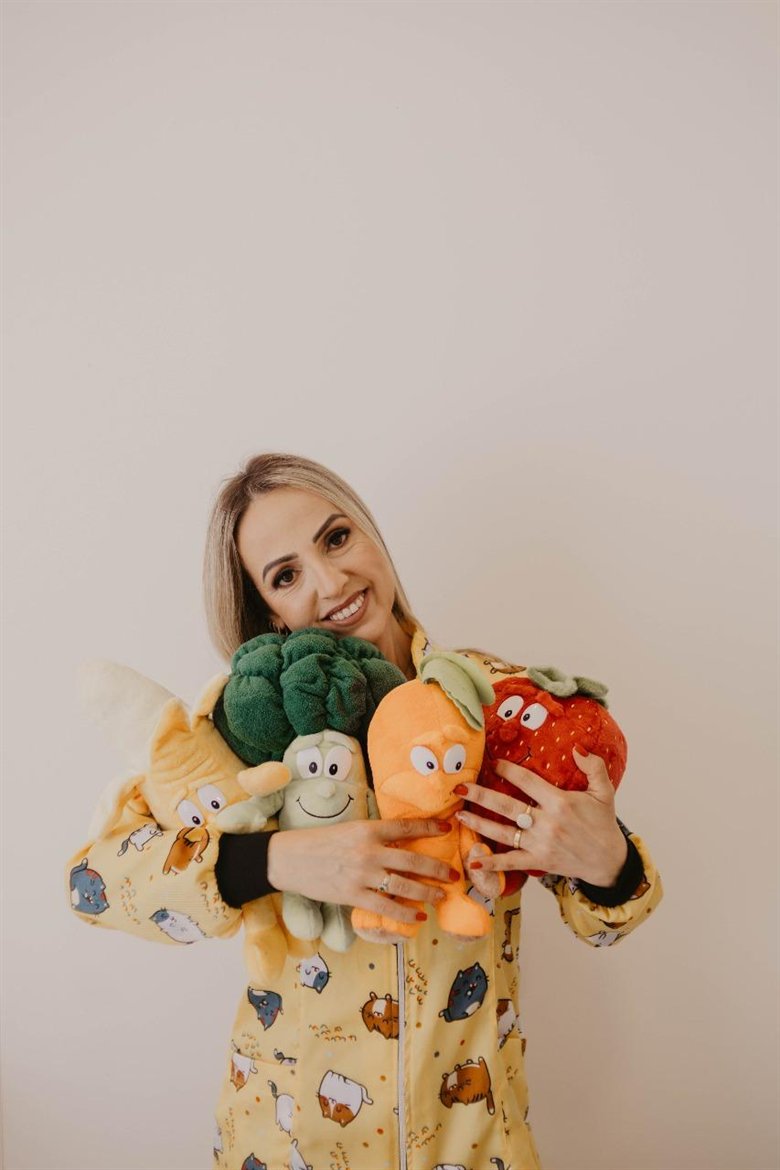 Rose Sodré, nutricionista, de jaleco amarelo, abraçando pelúcias de brócolis, cenoura, morango e banana